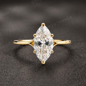 Puede incluir: Un anillo de oro con un diamante talla marquesa. El diamante está engastado en una montura de cuatro garras y la banda es de oro pulido. El anillo se muestra sobre una superficie gris oscuro.