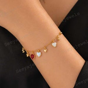 Può includere: Bracciale dorato con ciondoli a forma di cuore e pietre preziose. Il bracciale presenta un piccolo ciondolo iniziale, una gemma rossa, cuori bianchi opalini e piccoli cuori dorati.