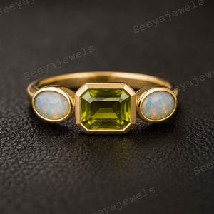 May include: A gold ring with a central emerald-cut green gemstone and two oval white gemstones. The ring is set against a dark backdrop, accentuating the gold band and the gemstones' sparkle. A stylish piece of jewellery.