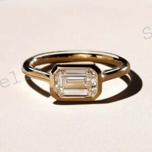 May include: A gold ring featuring a rectangular, clear gemstone. The gemstone is set within a bezel setting, and the band is plain, polished gold. The ring is presented on a light-coloured surface, with a shadow.