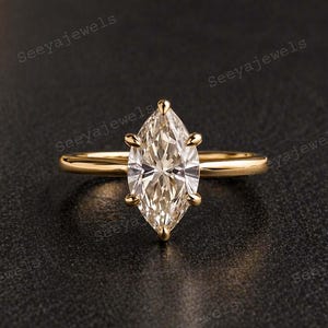 Puede incluir: Un anillo de oro con un diamante talla marquesa. El diamante está engastado con cuatro garras y tiene un corte brillante. La banda es delgada y pulida. El anillo se muestra sobre un fondo oscuro.