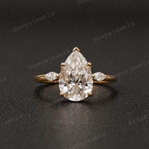 Puede incluir: Un anillo de oro con un gran diamante en forma de pera y diamantes más pequeños en forma de marquesa. El anillo se presenta sobre un fondo oscuro, resaltando el brillo de las piedras preciosas. El texto "Seeyajewels" es visible.