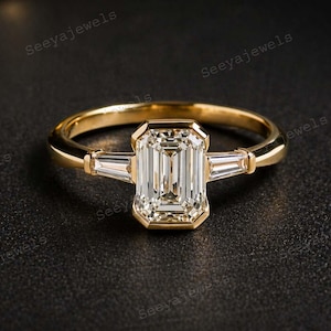May include: A gold ring with a large, rectangular, clear gemstone as the centerpiece. Two smaller, rectangular gemstones flank the main stone. The ring is set against a dark background.