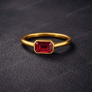 May include: A gold ring with a rectangular, deep red gemstone. The ring's band is thin and polished, and the gemstone is set within a gold bezel. The ring is displayed on a dark, textured surface.
