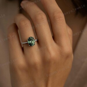 May include: An oval-cut green gemstone ring with a silver band, set with small accent stones. The ring is on a person's finger, showcasing the jewelry's design and color. The ring is a piece of fine jewelry.