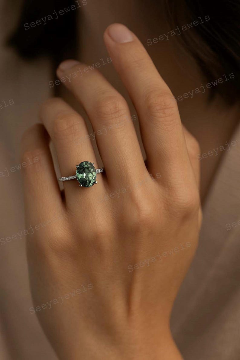 May include: An oval-cut green gemstone ring with a silver band, set with small accent stones. The ring is on a person's finger, showcasing the jewelry's design and color. The ring is a piece of fine jewelry.
