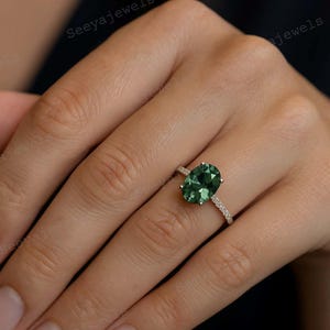 May include: An oval-cut, emerald-green gemstone ring with a delicate band of small, clear stones. The ring is set in a gold-colored metal and is displayed on a hand.