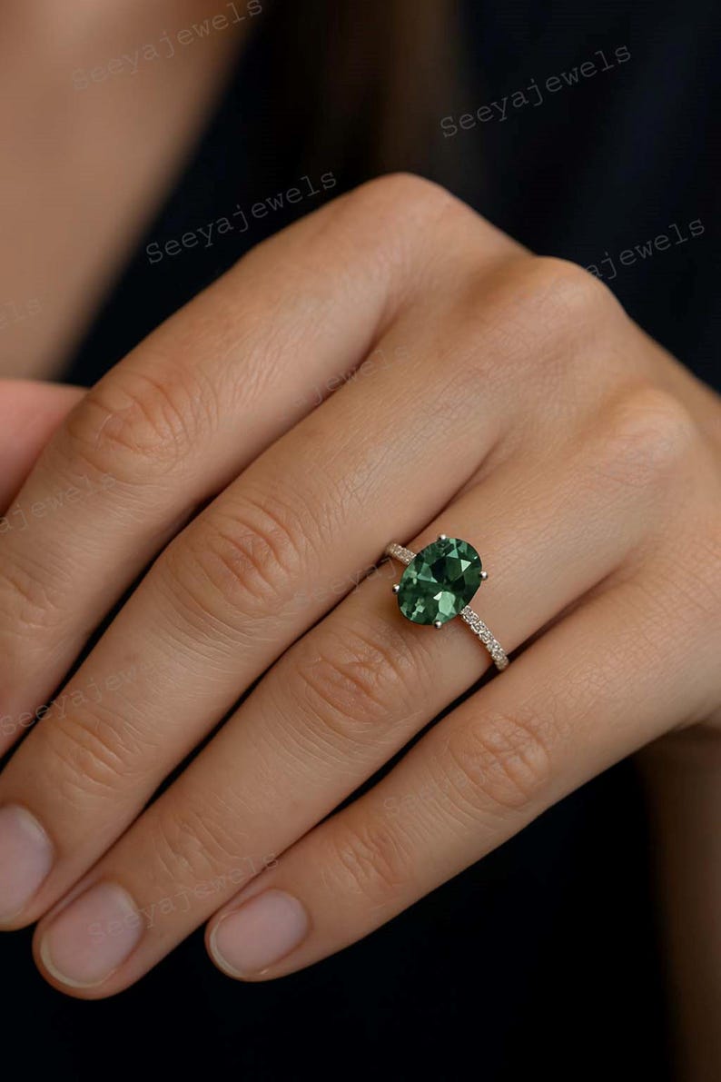 May include: An oval-cut, emerald-green gemstone ring with a delicate band of small, clear stones. The ring is set in a gold-colored metal and is displayed on a hand.