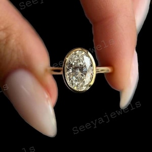 Könnte beinhalten: Ein goldener Ring mit einem großen, oval geschliffenen Diamanten. Der Diamant ist in einer Zargenfassung gefasst, und das Band ist dünn und filigran. Der Ring wird zwischen zwei Fingern vor schwarzem Hintergrund gehalten. Der Text "Seeyajewels" ist sichtbar.