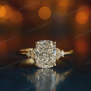 May include: A gold ring featuring a large, rectangular, clear gemstone. Smaller gemstones are set either side of the main stone. The ring is set against a blurred, warm-toned background. The text "Seeyajewels" is visible in the image.