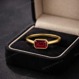 May include: A gold ring with a rectangular, deep red gemstone set in a bezel setting. The ring is displayed in a black jewelry box with a white trim. The brand name "Seeyajewels" is visible in the image.
