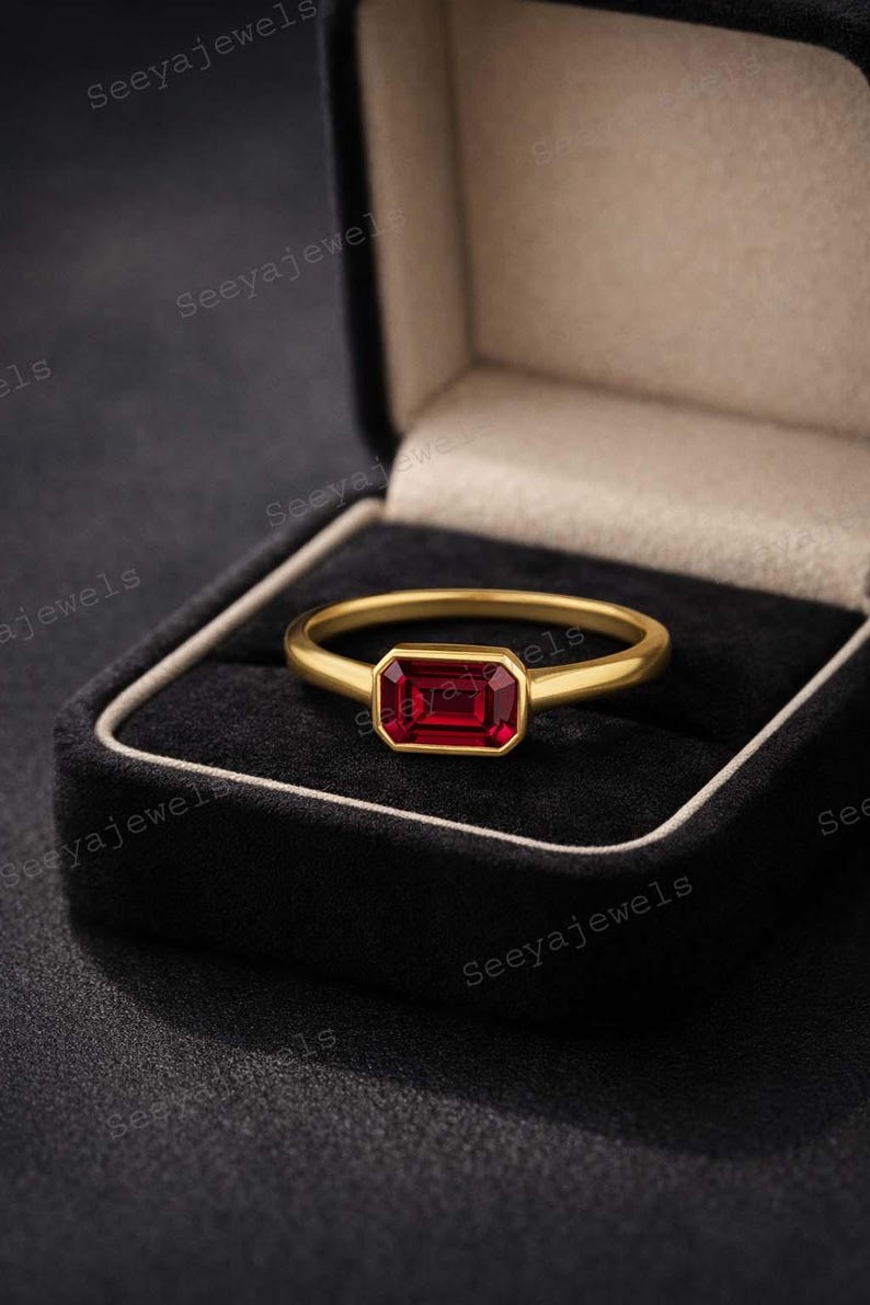 May include: A gold ring with a rectangular, deep red gemstone set in a bezel setting. The ring is displayed in a black jewelry box with a white trim. The brand name "Seeyajewels" is visible in the image.