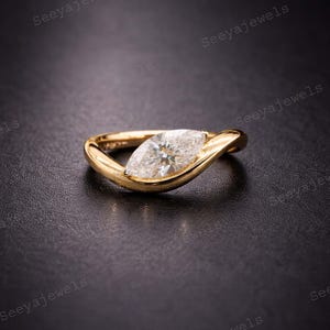 Puede incluir: Un anillo de oro con un diamante talla marquesa. El diamante está engastado en una banda curva y fluida. El anillo se encuentra sobre una superficie oscura, resaltando su diseño.