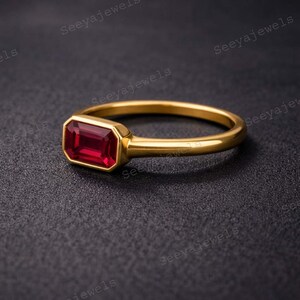 May include: A gold ring with a rectangular, deep red gemstone. The ring has a simple band and a bezel setting that holds the gemstone securely. The ring is displayed on a dark surface.