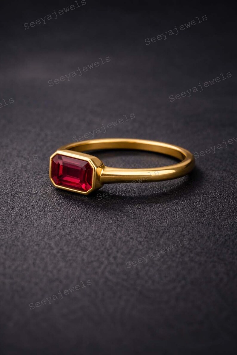 May include: A gold ring with a rectangular, deep red gemstone. The ring has a simple band and a bezel setting that holds the gemstone securely. The ring is displayed on a dark surface.