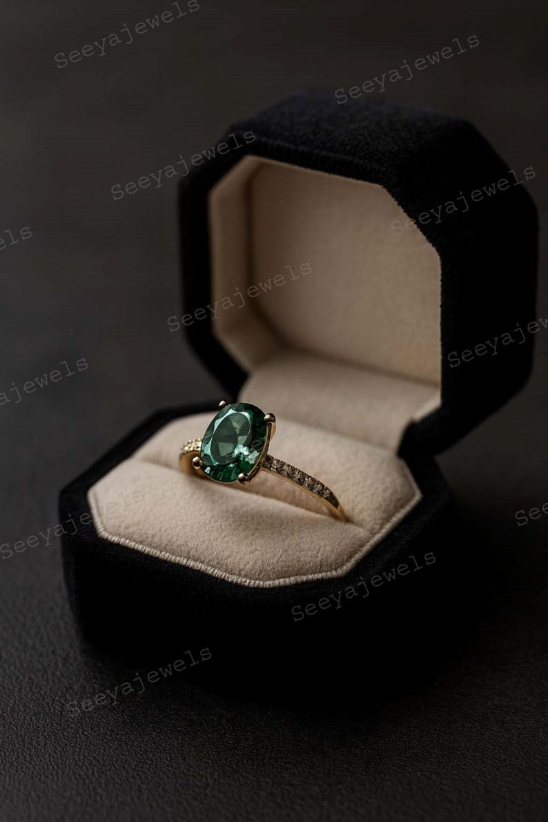May include: A gold ring with a large oval green gemstone, set in a black velvet jewelry box. The ring's band is adorned with small, sparkling stones. The box is open, revealing the ring against a beige interior.