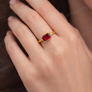 May include: A gold ring featuring a rectangular, deep red gemstone. The ring's band is a simple, polished gold. The gemstone is set in a bezel setting, showcasing its rich color. This piece is a classic jewelry item.
