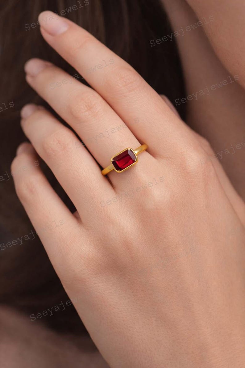 May include: A gold ring featuring a rectangular, deep red gemstone. The ring's band is a simple, polished gold. The gemstone is set in a bezel setting, showcasing its rich color. This piece is a classic jewelry item.