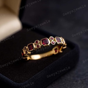 Op de afbeelding: Een gouden ring met afwisselend vierkante en ronde rode edelstenen. De ring is gezet in een gouden band en wordt gepresenteerd in een zwarte sieradendoos. De ring is een sieraad.