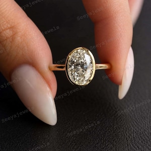 May include: A gold ring with an oval-cut diamond. The diamond is set in a bezel setting. The ring is held up by a person's hand. The ring is a piece of jewelry.