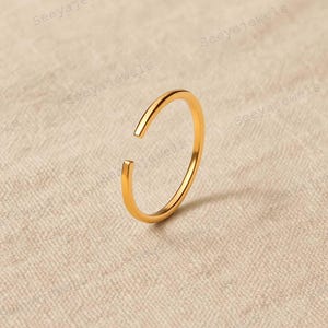 May include: A gold-coloured open ring. The ring has a simple, minimalist design with a gap at the top. The ring is made of a shiny, reflective metal. The ring is shown on a neutral-coloured background.