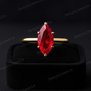 May include: A gold ring featuring a marquise-cut, vibrant red gemstone. The ring is set in a gold band and is displayed on a black surface. The gemstone is the focal point, showcasing its rich color and shape. The ring is a piece of jewelry.