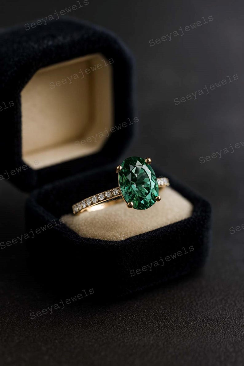 May include: An emerald-cut green gemstone ring with a gold band, set in a black velvet ring box. The band is adorned with small, sparkling diamonds. The ring is a piece of fine jewelry.