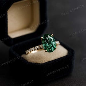 May include: An emerald-cut green gemstone ring with a gold band, set in a black velvet ring box. The band is adorned with small, sparkling diamonds. The ring is a piece of fine jewelry.