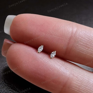 May include: Two small, gold-colored stud earrings, each featuring a marquise-cut, clear gemstone. The earrings are set on a finger, showcasing their delicate size and design. The background is a blurred view of the finger.