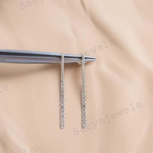 May include: A pair of silver-toned, linear drop earrings. Each earring is a slender, vertical bar encrusted with small, sparkling stones. The earrings are held by a silver tool against a neutral background, showcasing their delicate design.