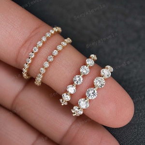 Puede incluir: Cuatro anillos dorados con piedras preciosas redondas y transparentes. Dos anillos tienen piedras más pequeñas, mientras que los otros dos tienen gemas más grandes. Los anillos se muestran en un dedo sobre un fondo oscuro.
