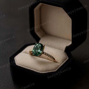 May include: A gold ring with a large oval green gemstone, set in a black velvet jewelry box. The ring's band is adorned with small, sparkling stones. The box is open, revealing the ring against a beige interior.