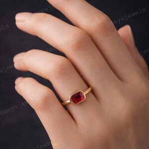 May include: A gold ring with a rectangular, deep red gemstone. The ring is set in a simple bezel setting, showcasing the vibrant color of the stone. The ring is on a hand.
