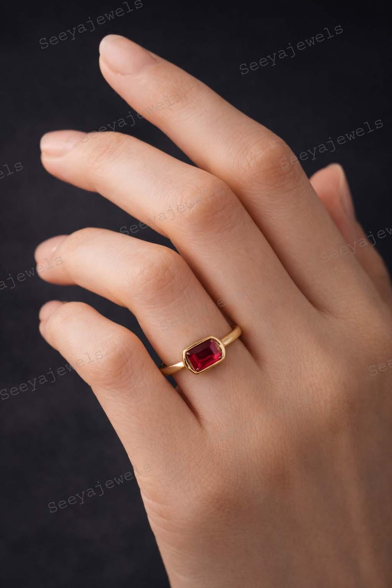 May include: A gold ring with a rectangular, deep red gemstone. The ring is set in a simple bezel setting, showcasing the vibrant color of the stone. The ring is on a hand.