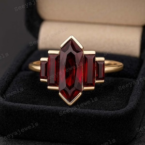 Op de afbeelding: Een gouden ring met een centrale, langwerpige, dieprode edelsteen, geflankeerd door twee kleinere rechthoekige edelstenen aan elke kant. De ring is gezet in een gouden band en wordt gepresenteerd in een zwarte sieradendoos. Het ontwerp van de ring is geometrisch en geïnspireerd op Art Deco.