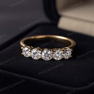 Könnte beinhalten: Ein goldener Ring mit fünf runden, klaren Edelsteinen. Der Ring ist vor einem schwarzen Hintergrund platziert, der das Funkeln der Steine hervorhebt. Ein zeitloses Schmuckstück für verschiedene Anlässe.