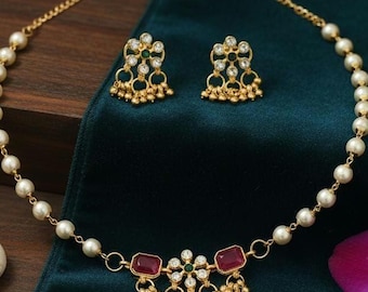 May include: A gold-toned jewelry set featuring a necklace and matching earrings. The necklace has a pearl strand with a pendant of red and clear stones. The earrings have a similar design with green and clear stones. The set is displayed on a dark green fabric.
