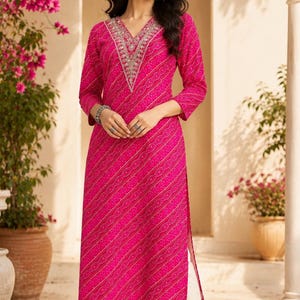 May include: A vibrant fuchsia pink straight-cut kurta featuring a white geometric bandhani-style pattern, three-quarter sleeves, and a V-neckline with intricate silver embroidery. The garment is paired with plain off-white straight-leg trousers.