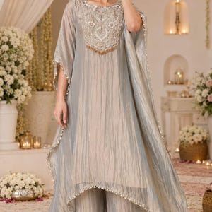 May include: A light gray kaftan-style tunic featuring a textured, shimmering fabric with a high-low hemline and intricate silver embroidery around the neckline. The garment is paired with matching wide-leg trousers and finished with delicate beaded trim.