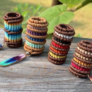 May include: Four stacked, round, brown macrame hair ties in various colors, including blue, purple, red, and yellow. Each tie has a small, decorative feather charm attached. The ties are arranged on a weathered wooden surface.