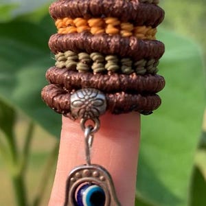 Könnte beinhalten: Ein gestapelter Makramee-Ring in Braun-, Türkis- und Gelbtönen, mit einem silbernen Anhänger in Form einer Hamsa-Hand mit einem bösen Blick. Der Ring wird an einem Finger getragen und zeigt die komplizierte Knotenarbeit.