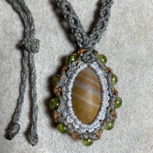 May include: A handmade macrame necklace featuring a central oval stone with brown and tan stripes. The stone is surrounded by a gray and brown macrame design, accented with green beads. The necklace has a gray cord.