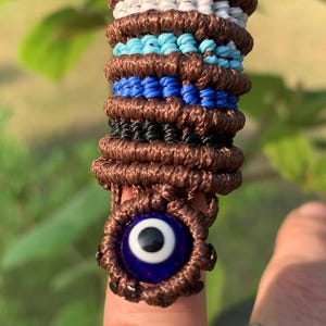 May include: A macrame ring featuring a brown, white, and blue design with an evil eye charm. The ring is made of woven cord and has a circular evil eye with a blue iris and white pupil.
