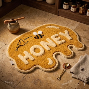 Puede incluir: Una alfombra amarilla con forma de derrame de miel con la palabra "HONEY" en letras color crema. Una abeja de dibujos animados y un corazón están en la alfombra. A la izquierda hay un pequeño tarro de miel de cerámica con un cucharón de miel de madera.