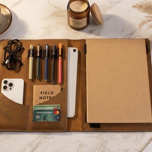 May include: A brown leather folio organizer with multiple compartments. It holds a notepad, pens, a smartphone, a tablet, and a small notebook labeled "FIELD NOTES." The folio also has slots for cards and a charging cable. A cup of coffee and a candle are in the background.