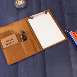 Op de afbeelding: Open, bruin lederen clipboard met een zilveren clip met een blanco wit papier. Een zakje bevat een klein notitieboekje met de tekst "FIELD NOTES" en twee pennen. Een set kleurpotloden en een kandelaar zijn ook zichtbaar.