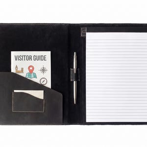 May include: A black leather portfolio featuring a white lined notepad on the right side, a silver pen held in a center loop, and a left-side pocket containing a white booklet with the text VISITOR GUIDE and colorful map icons printed on it.