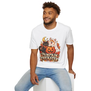 Może przedstawiać: Biały t-shirt z nadrukiem graficznym przedstawiającym czarnego kota i psa w przebraniu ducha siedzących za dynią. Wzór zawiera jesienne liście, półksiężyc oraz napis PUMPKIN TRICKSTER wykonany stylizowaną czcionką.