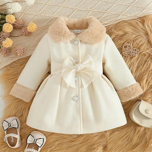 May include: Cream-colored toddler's coat with a faux fur collar and cuffs. The coat features a large bow at the waist and decorative buttons. Two pairs of white and black bow-adorned shoes are at the bottom of the image.
