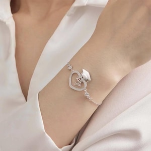 May include: A silver bracelet with a heart-shaped charm inscribed with "2024" and "Jadie", and a graduation cap. The bracelet has small, sparkling details. A thoughtful graduation gift.